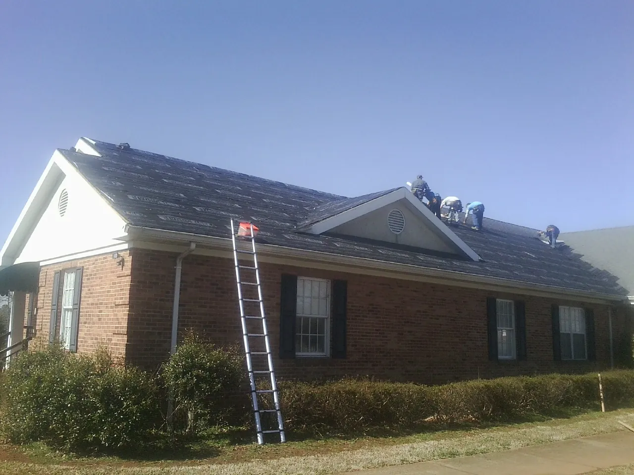 Crew tearing shingles off a brick ranch roof with a ladder set — Emergency Roof Repair work by our crew in Wixom