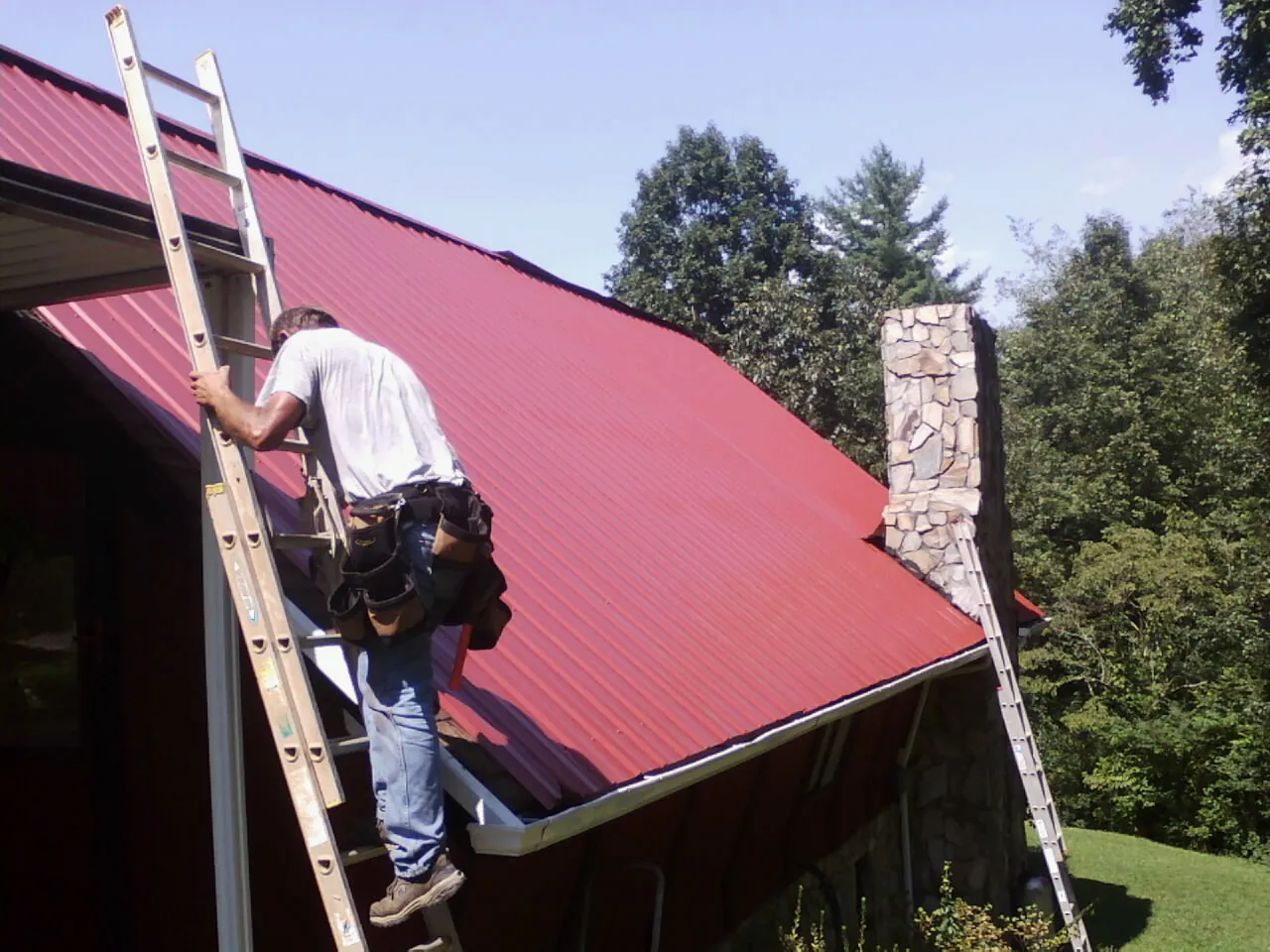 Roofer on a ladder beside a red metal roof and stone chimney — Soffit & Fascia Repair by Roofing Brothers in Wixom